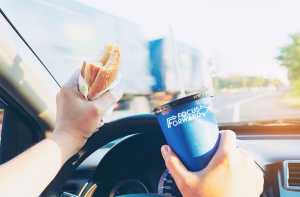 pictured is a driver gripping a burger and soda while driving a moving car