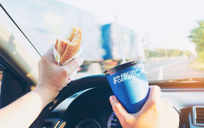 pictured is a driver gripping a burger and soda while driving a moving car